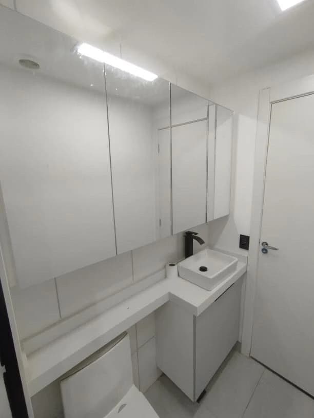 Banheiro Minimalista e Funcional: Ambiente em tons de branco, com móveis em MDF e bancada de apoio. Destaque para os espelhos amplos e iluminação integrada, promovendo uma sensação de amplitude.
